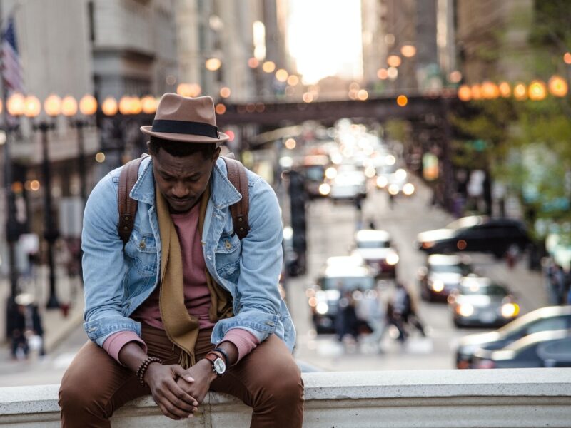A man in a leather jacket looking down while sitting on a ledge in a city